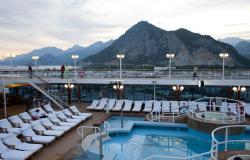 Azamara Journey - Azamara Club Cruises - rozsvícené lampy na horní palubě lodi a panorama skalnatých vrcholků na pobřeží