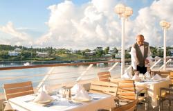 Azamara Journey - Azamara Club Cruises - čísník připravujíc jídelní stoly na venkovní terasa a v pozadí panorama luxusních vil a domků na zeleném pobřeží